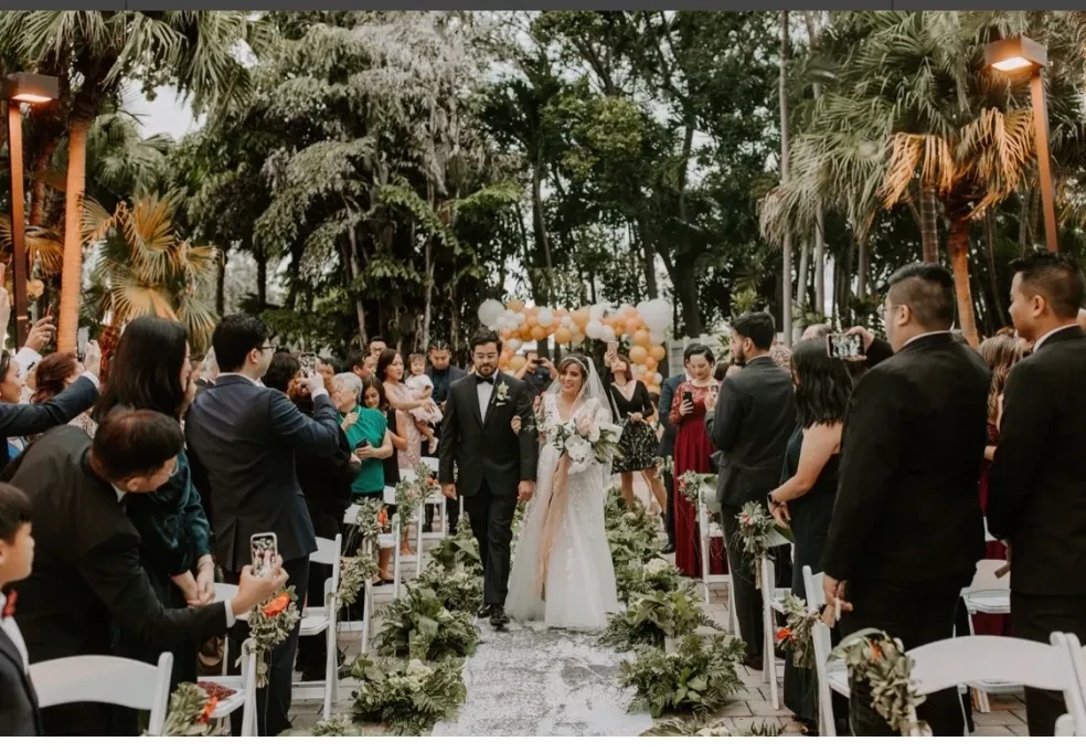 Couple walking down aisle lined with greenery and white chairs