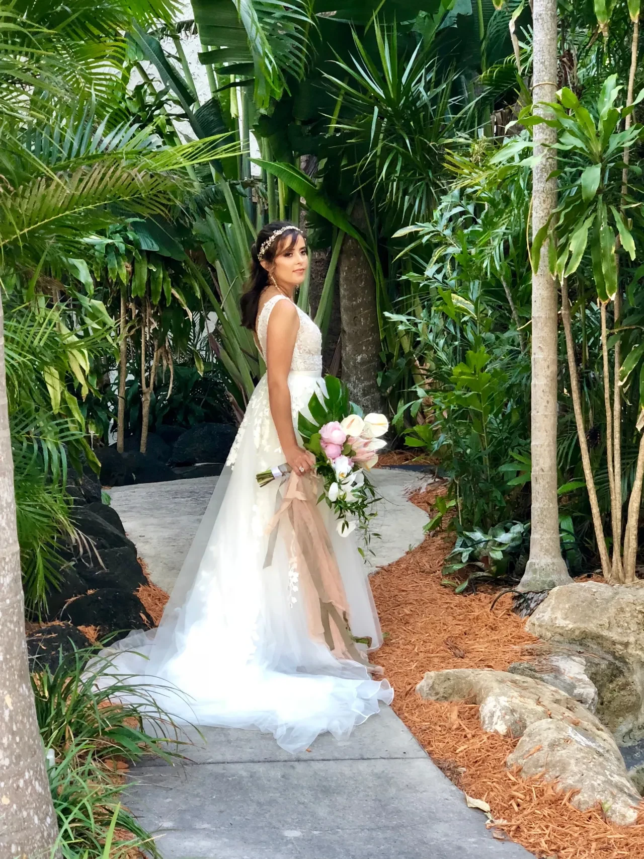 Bride in white gown with pink bouquet in tropical garden path
